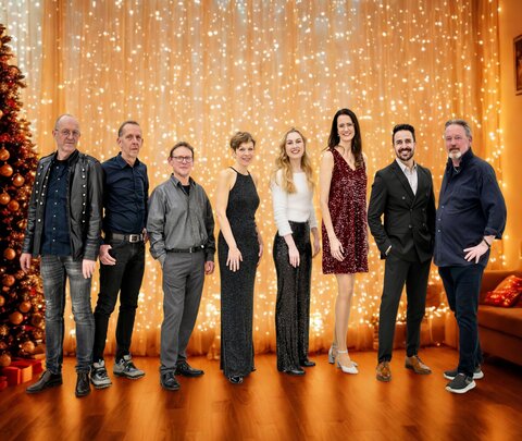 Gruppenfoto von acht Personen in festlicher Kleidung vor einem warm beleuchteten Hintergrund mit goldenem Lichtervorhang und weihnachtlich geschmücktem Baum. Die Personen stehen nebeneinander und lächeln in die Kamera.