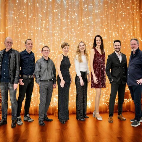 Gruppenfoto von acht Personen in festlicher Kleidung vor einem warm beleuchteten Hintergrund mit goldenem Lichtervorhang und weihnachtlich geschmücktem Baum. Die Personen stehen nebeneinander und lächeln in die Kamera.