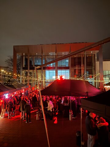 Nächtliche Veranstaltung mit zahlreichen Besuchern auf einem beleuchteten Platz vor einem modernen Glasgebäude. Schwarze Marktzelte, rote Lichtinstallationen und Lichterketten sorgen für eine festliche Atmosphäre.