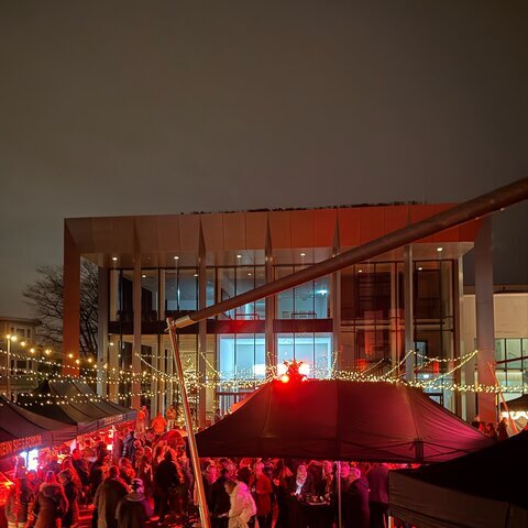 Nächtliche Veranstaltung mit zahlreichen Besuchern auf einem beleuchteten Platz vor einem modernen Glasgebäude. Schwarze Marktzelte, rote Lichtinstallationen und Lichterketten sorgen für eine festliche Atmosphäre.