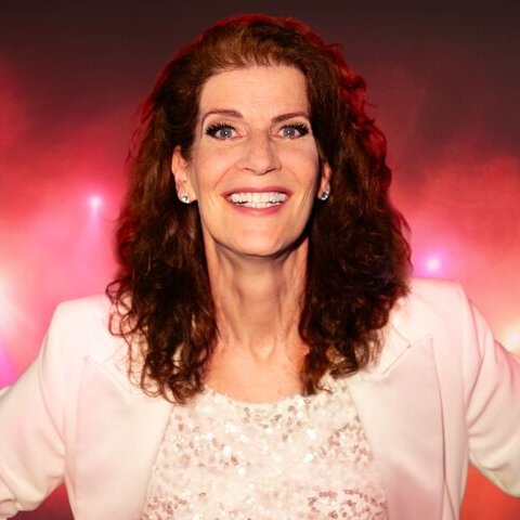 Porträt einer lächelnden Frau mit dunkelbraunen, lockigen Haaren, hellem Blazer und glitzerndem Oberteil vor einem rot-pink beleuchteten Hintergrund.