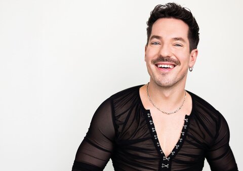 Künstler Niko Stank in schwarzem Hemd lachend vor weißem Hintergrund