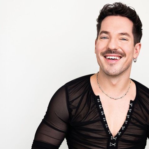 Künstler Niko Stank in schwarzem Hemd lachend vor weißem Hintergrund