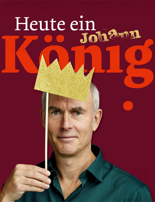 Porträt von Comedian Johann König vor dunkelrotem Hintergrund. Er hält eine goldene Papierkrone vor sein Gesicht. Über dem Bild steht in großer Schrift „Heute ein König“ mit seinem Namen „Johann“ darüber.
