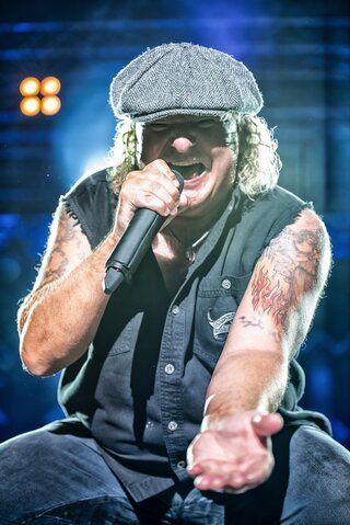 Nahaufnahme eines Rocksängers bei einem Live-Konzert. Mit Schiebermütze, ärmelloser schwarzer Weste und sichtbaren Arm-Tattoos singt er kraftvoll in ein Mikrofon und streckt die Hand dem Publikum entgegen. Bühnenlicht und unscharfe Scheinwerfer im Hintergrund unterstreichen die energiegeladene Konzertatmosphäre.