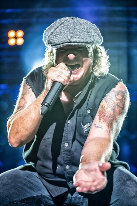 Nahaufnahme eines Rocksängers bei einem Live-Konzert. Mit Schiebermütze, ärmelloser schwarzer Weste und sichtbaren Arm-Tattoos singt er kraftvoll in ein Mikrofon und streckt die Hand dem Publikum entgegen. Bühnenlicht und unscharfe Scheinwerfer im Hintergrund unterstreichen die energiegeladene Konzertatmosphäre. Nahaufnahme eines Rocksängers bei einem Live-Konzert. Mit Schiebermütze, ärmelloser schwarzer Weste und sichtbaren Arm-Tattoos singt er kraftvoll in ein Mikrofon und streckt die Hand dem Publikum entgegen. Bühnenlicht und unscharfe Scheinwerfer im Hintergrund unterstreichen die energiegeladene Konzertatmosphäre.