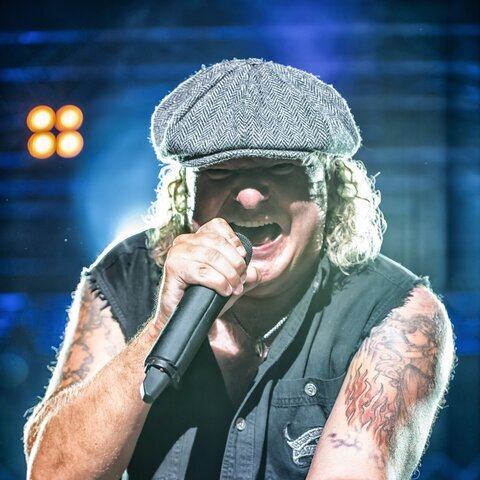 Nahaufnahme eines Rocksängers bei einem Live-Konzert. Mit Schiebermütze, ärmelloser schwarzer Weste und sichtbaren Arm-Tattoos singt er kraftvoll in ein Mikrofon und streckt die Hand dem Publikum entgegen. Bühnenlicht und unscharfe Scheinwerfer im Hintergrund unterstreichen die energiegeladene Konzertatmosphäre.