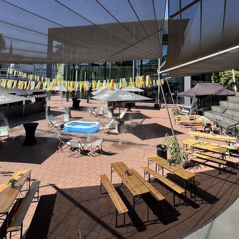 Festlich gestalteter Außenbereich des RHEIN SIEG FORUM mit Bierzeltgarnituren, Stehtischen und Sonnenschirmen. Bunte Bänder überspannen den Platz, in der Mitte steht ein kleines Planschbecken. Im Hintergrund ist der Eingangsbereich des Veranstaltungsortes zu sehen.